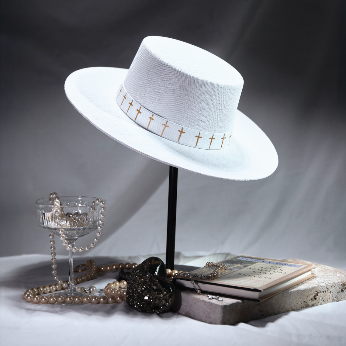 White hat with gold accents on a stand, surrounded by books and jewelry on a dark background.
